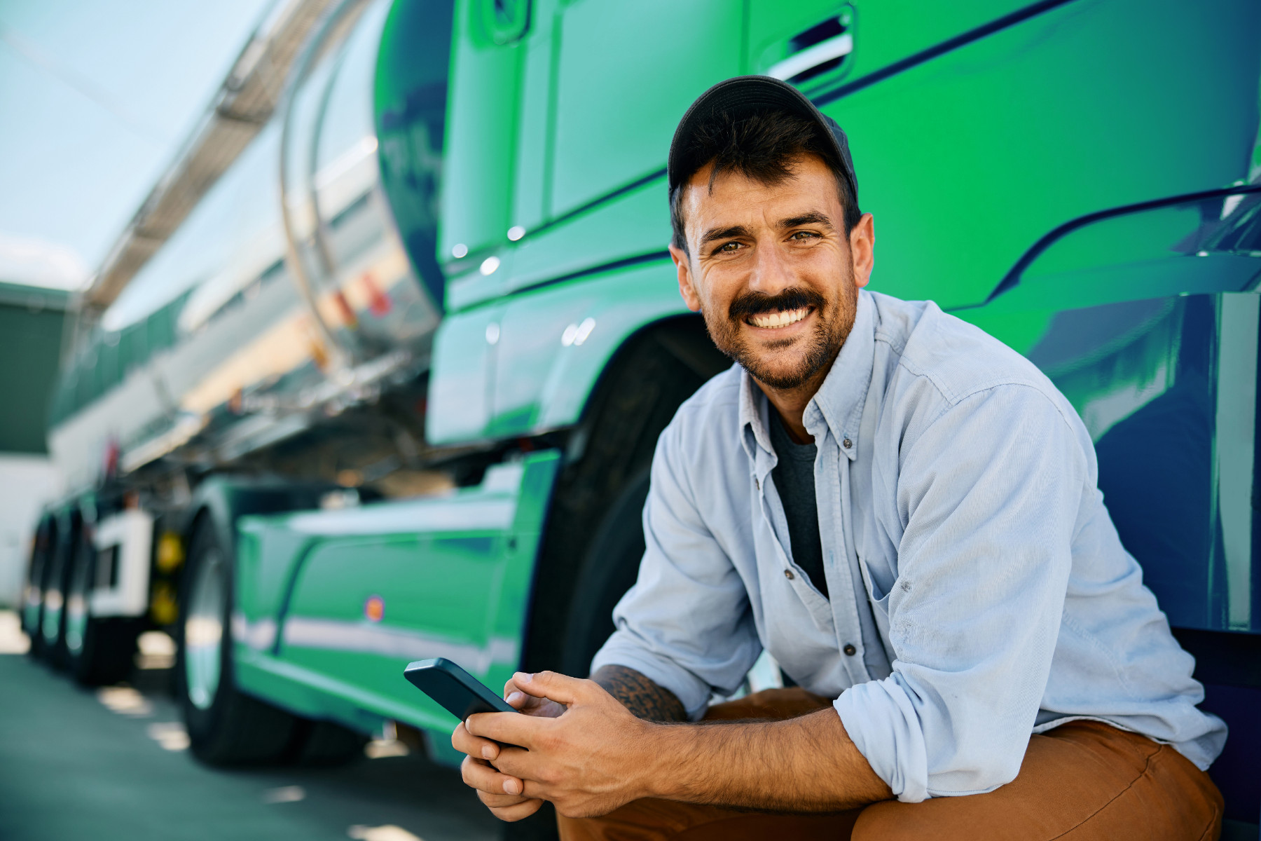 L&auml;chelnder Lkw-Fahrer mit Smartphone vor einem gr&uuml;nen Tanklaster
