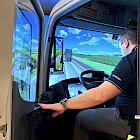 Person sitzt in einem DEKRA-Lkw-Fahrsimulator mit Stra&szlig;enansicht auf den Bildschirmen