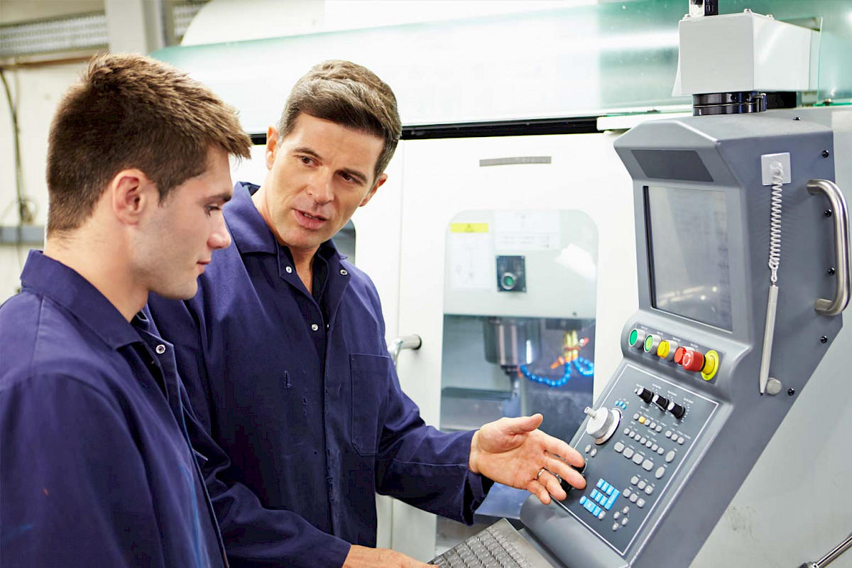 Facharbeiter weist einen jungen Auszubildenden an einer CNC-Maschine ein